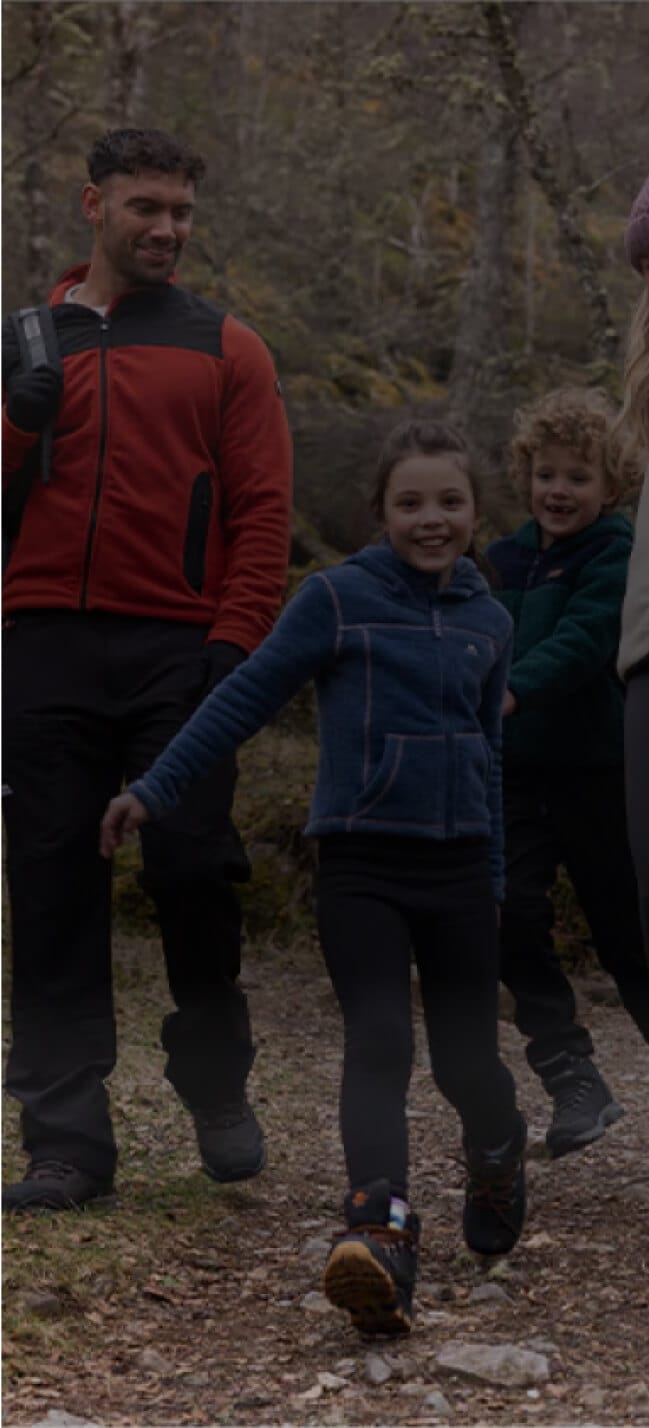 A man and two children stroll along a forest trail, centric in their enjoyment. The man, in a red jacket, smiles warmly at the children. The girl in a blue jacket and the boy in a green jacket cheerfully walk ahead, immersed in nature’s embrace.